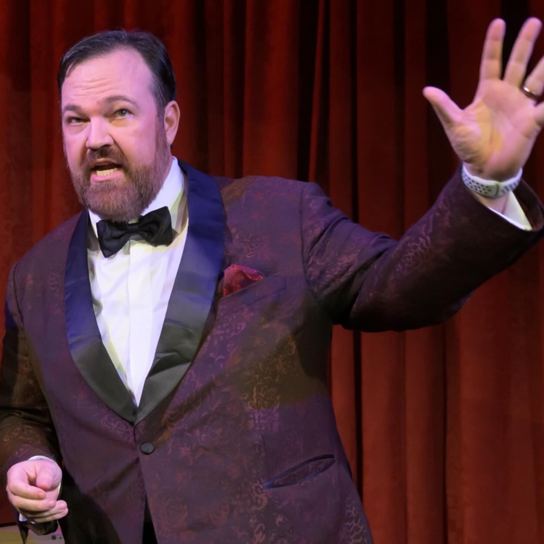 Matt Donnelly captivates the audience with his powerful stage presence, dressed in a sharp tuxedo against a rich red curtain backdrop.