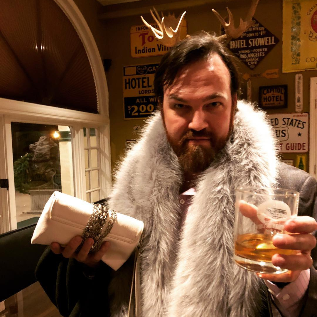 A regal moment captured—Matt Donnelly embraces a luxurious persona with a fur coat, a drink in hand, and an intense gaze.